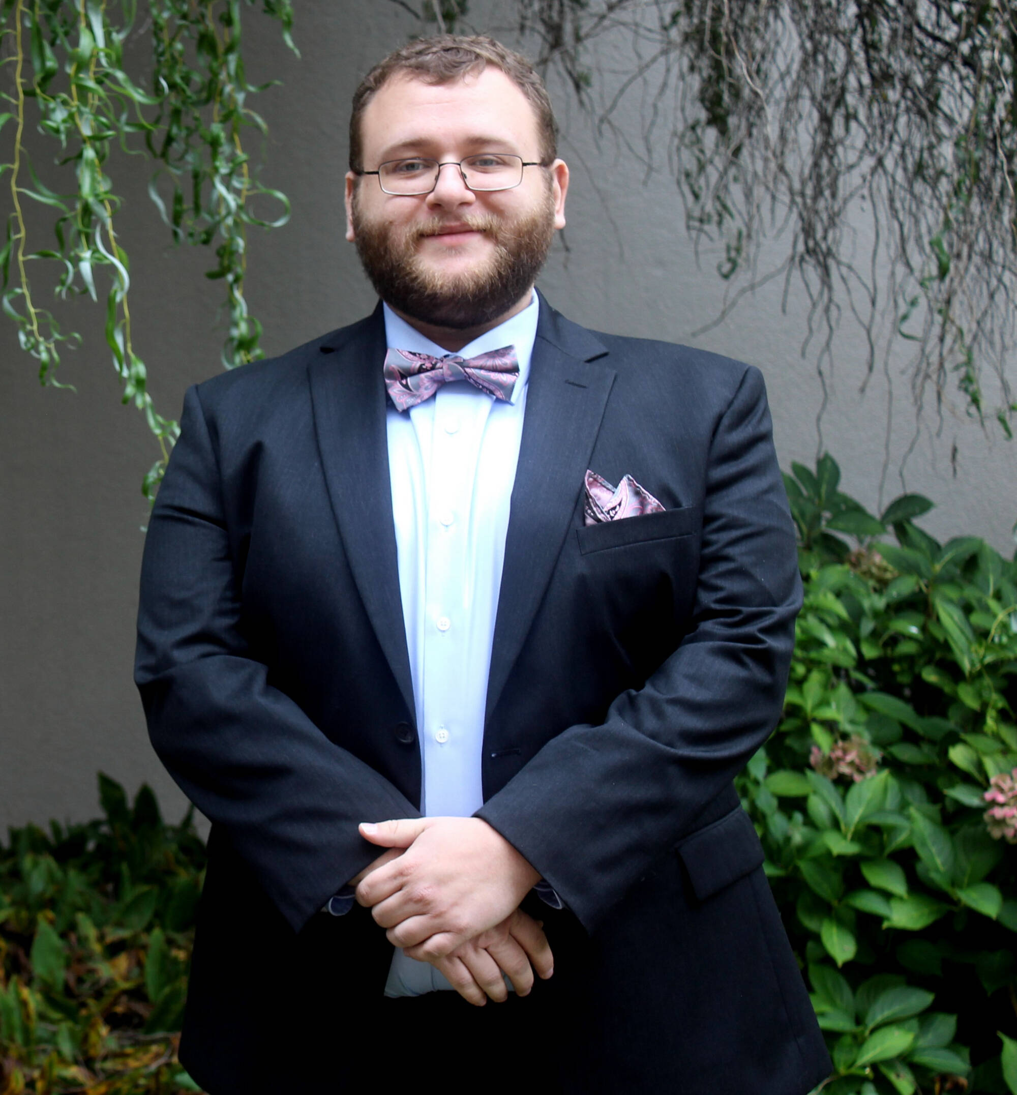 louis Marx is wearing a dark blue suit, white shirt, and a patterned bow tie with a matching pocket square, standing with hands clasped in front of a green leafy background